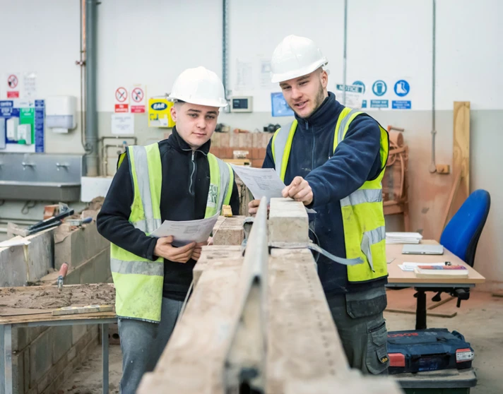 Two brickwork students in safety gear reviewing plans together, standing beside a brick wall and discussing their next steps in a practical training workshop. Two brickwork students in safety gear reviewing plans together, standing beside a brick wall and discussing their next steps in a practical training workshop.
